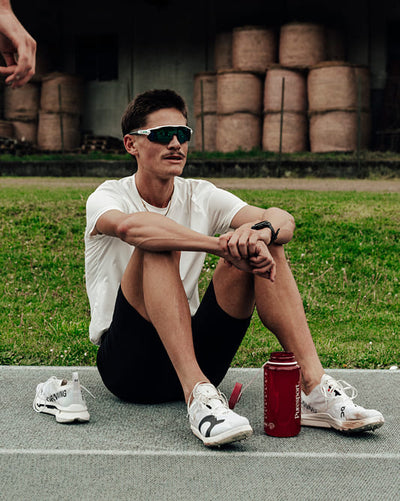 Man sitting on a concrete surface with sports equipment, wearing sunglasses and a white shirt.