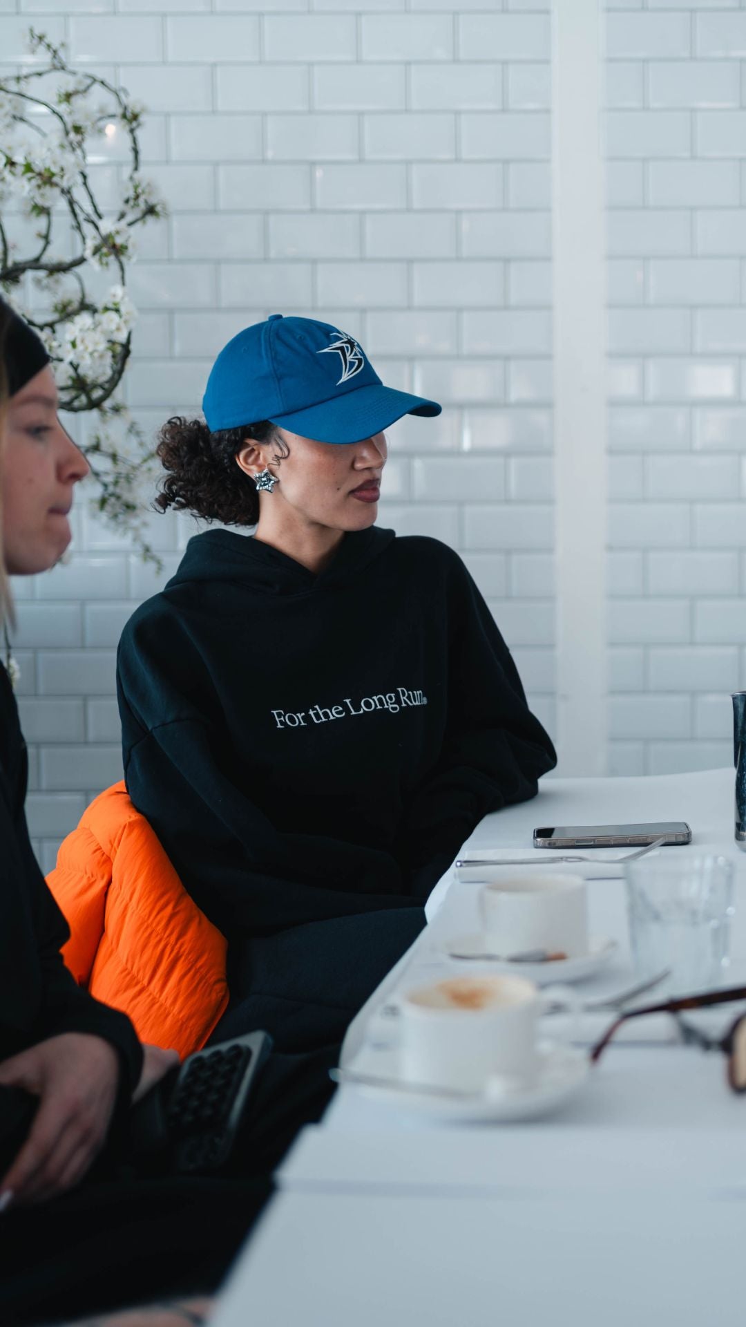 Woman wearing a blue cap and black Puresport hoodie with For the Long Run, sitting at a table with coffee cups.