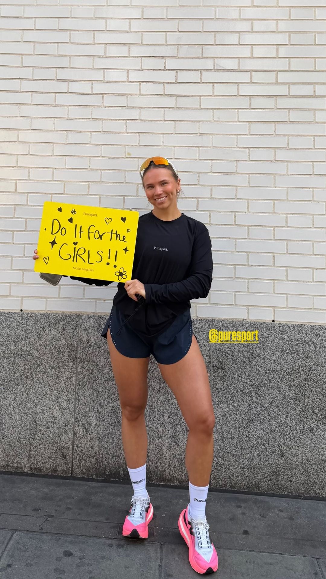 A women runner holding a yellow sign with Puresport branding at Hackney half, standing against a light gray brick wall.