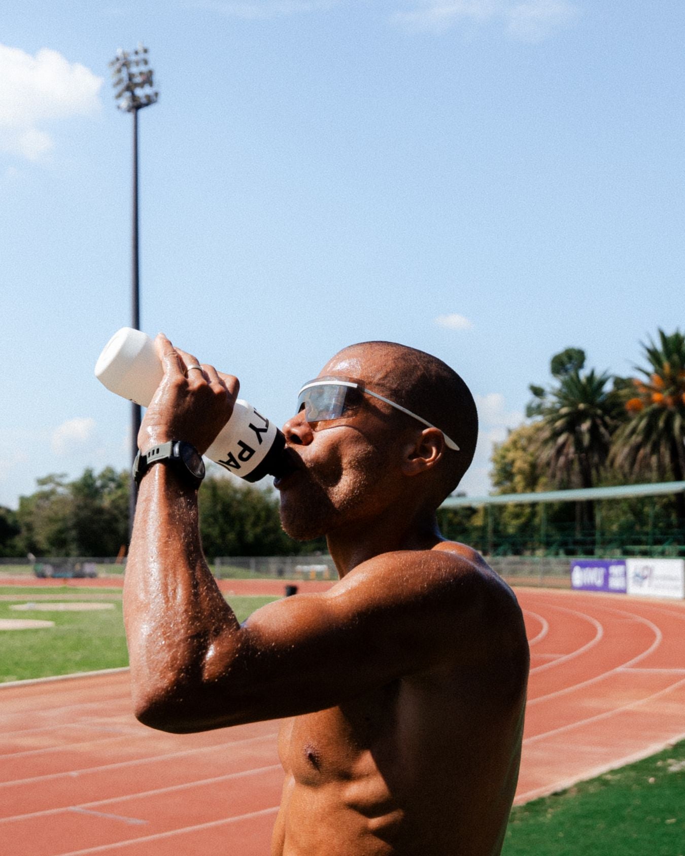 Elliot Giles drinking electrolytes from a water bottle on a running track with palm trees in the background