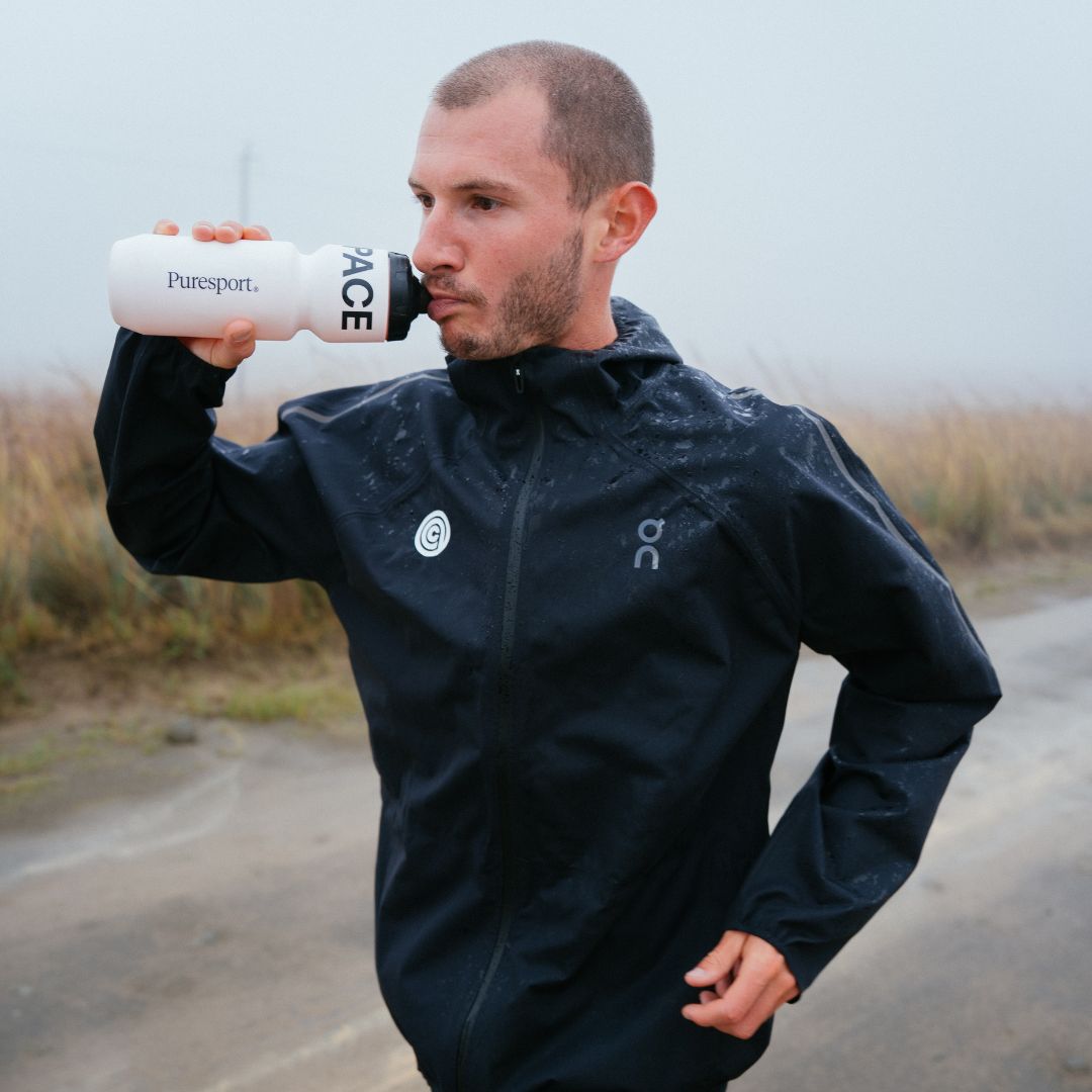 George Mills in a black jacket holding a white water bottle with 'Puresport' branding on a road.