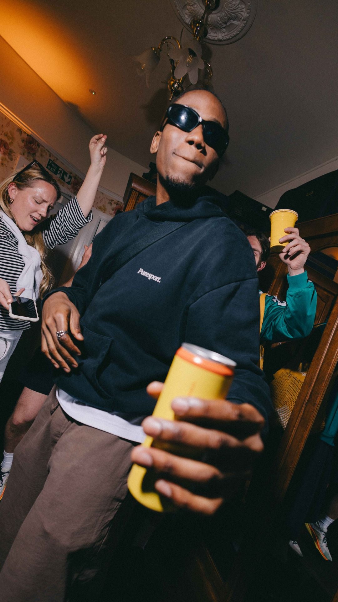 A person wearing sunglasses and a Puresport hoodie holding a yellow cup at House Party in Soho.