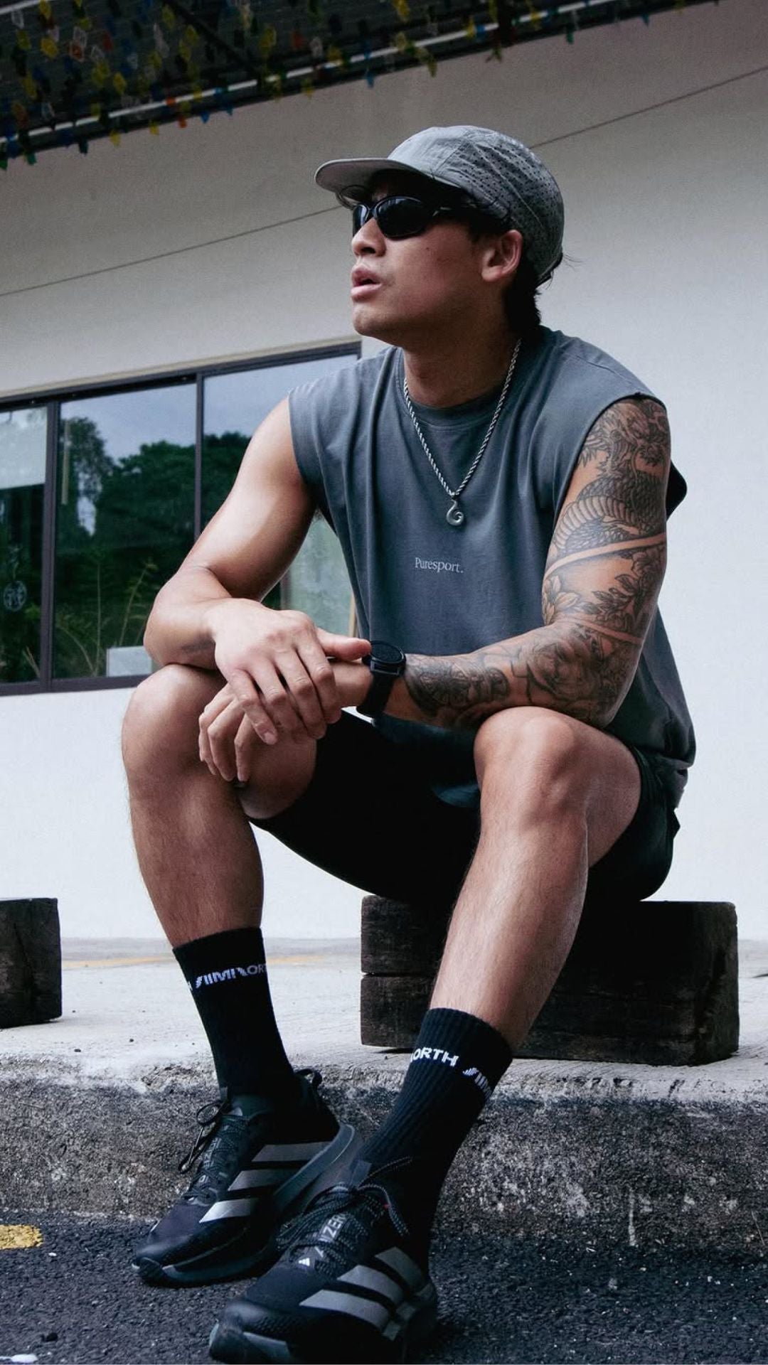 Man sitting on post wearing a sleeveless shirt by Puresport, cap, sunglasses, and black shoes.