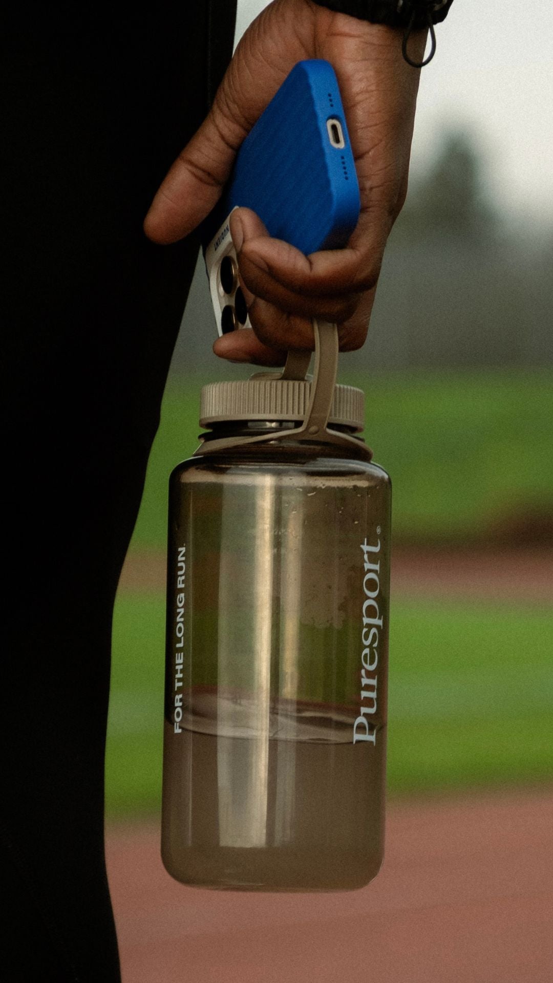 Person holding a clear nalgene bottle with a blue power bank attached, on a blurred outdoor background.
