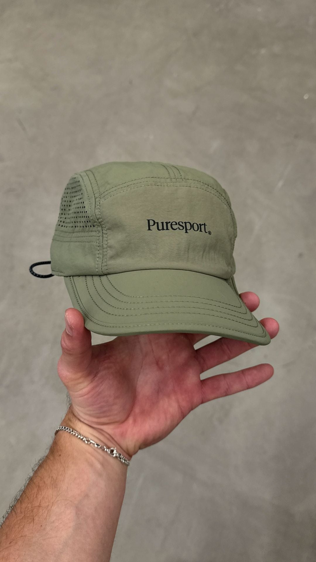 Olive cap with 'Puresport' logo held in a hand against a grey background