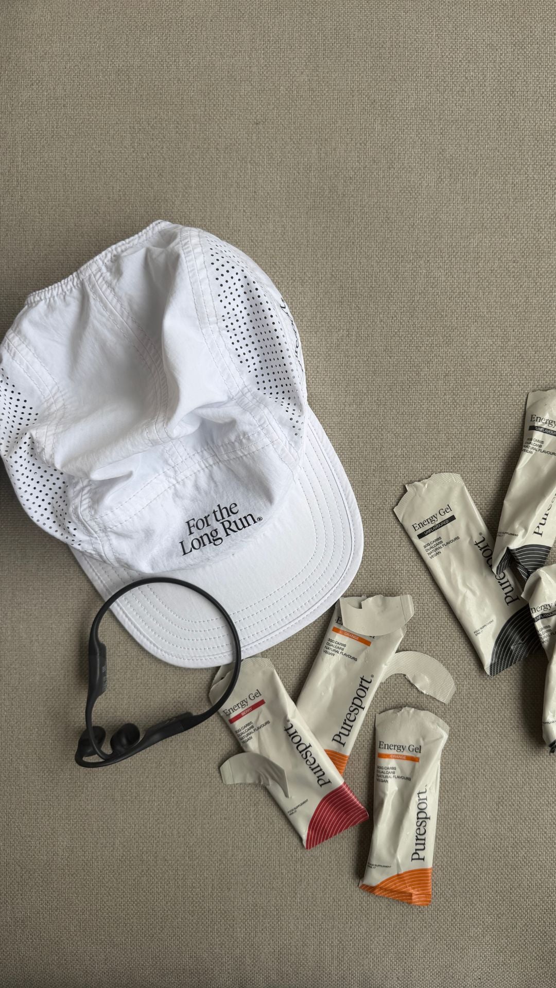 White cap with 'For the Long Run' text, earbuds, and energy gels on a gray surface
