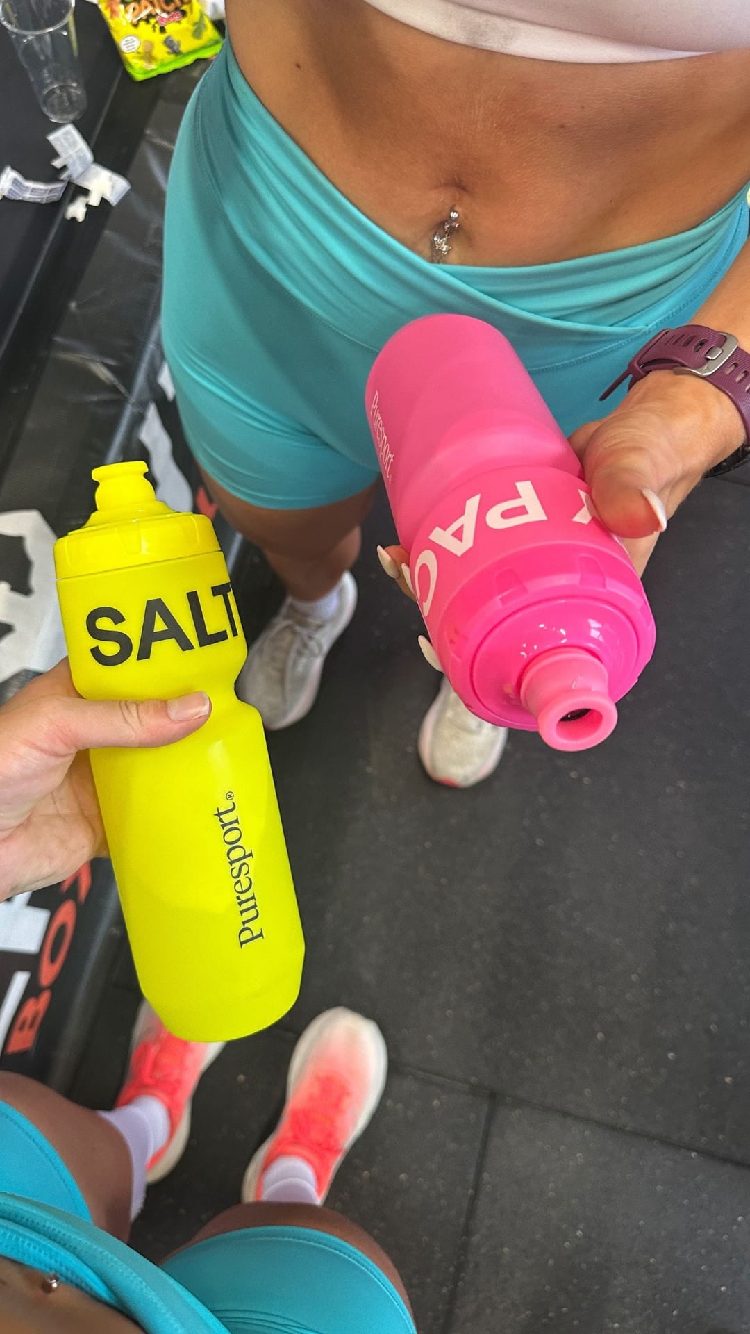 Two bottles, one yellow and one pink, held by two women during a hyrox session