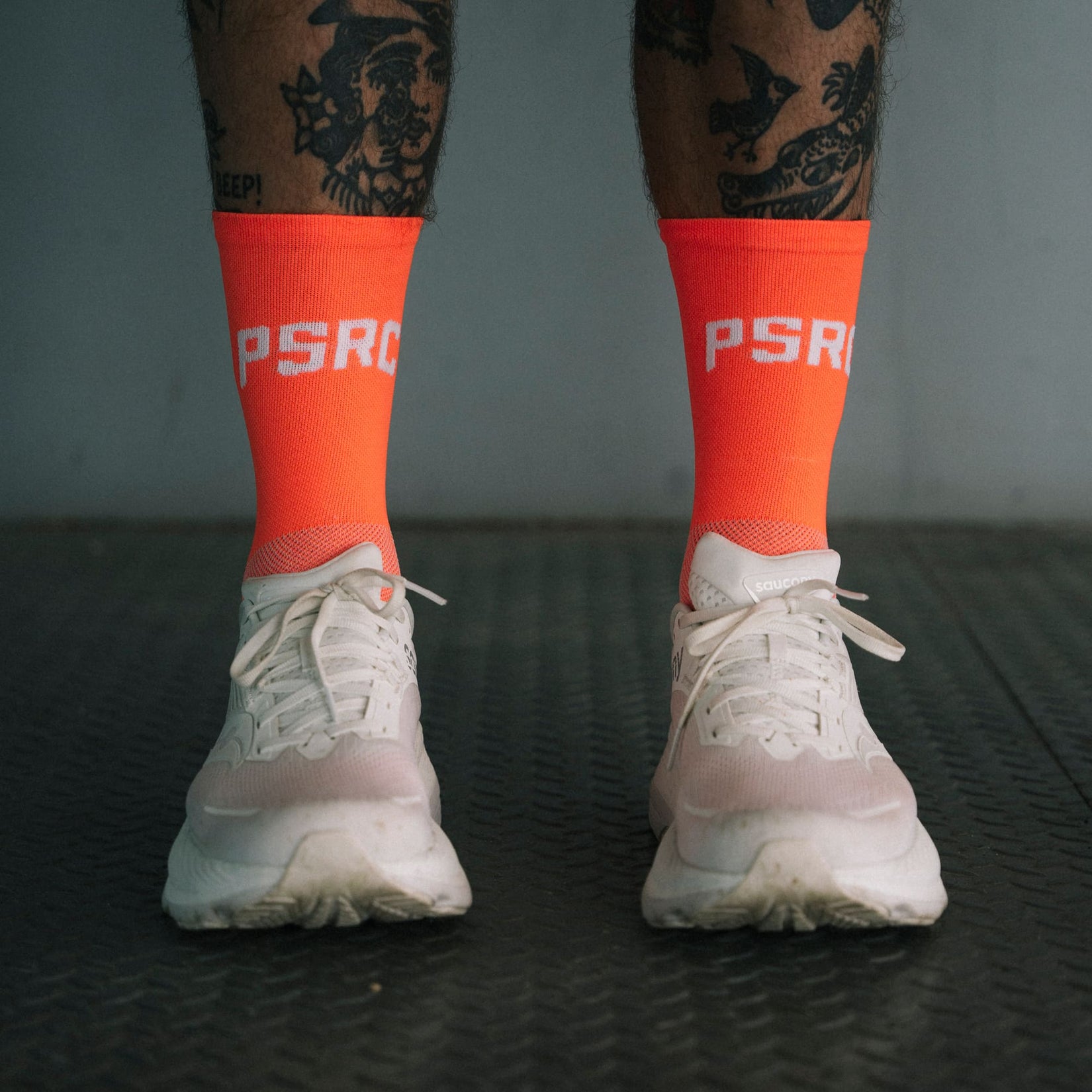 Person wearing orange socks with 'PSRC' branding and white sneakers on a dark textured floor.