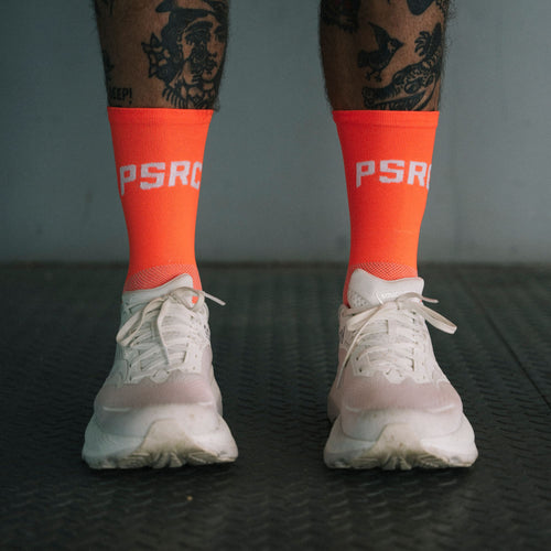 Person wearing orange socks with 'PSRC' branding and white sneakers on a dark textured floor.