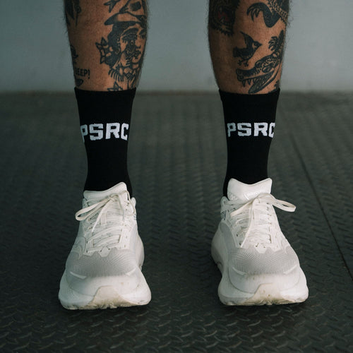 Person wearing white sneakers and black socks with 'PSRC' logo on a dark textured floor.