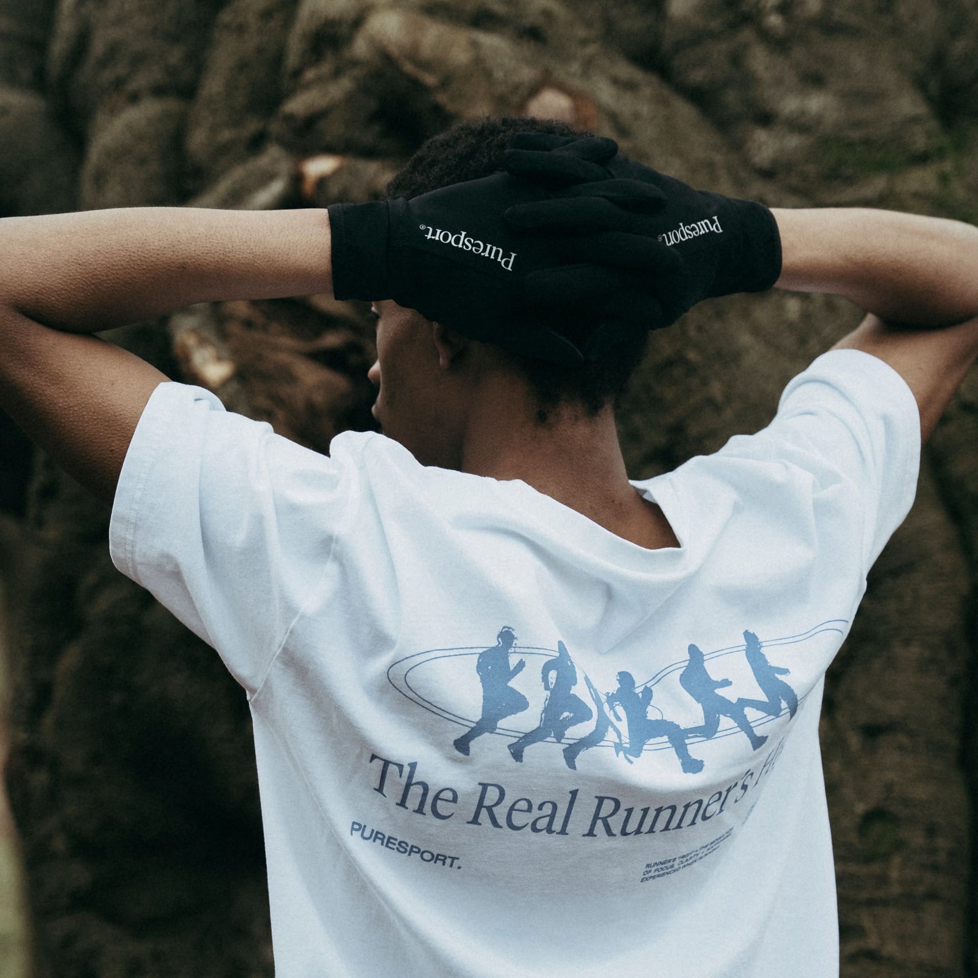 Person wearing a white pureport t-shirt with 'The Real Runners' design and black Puresport gloves, standing in a forest.