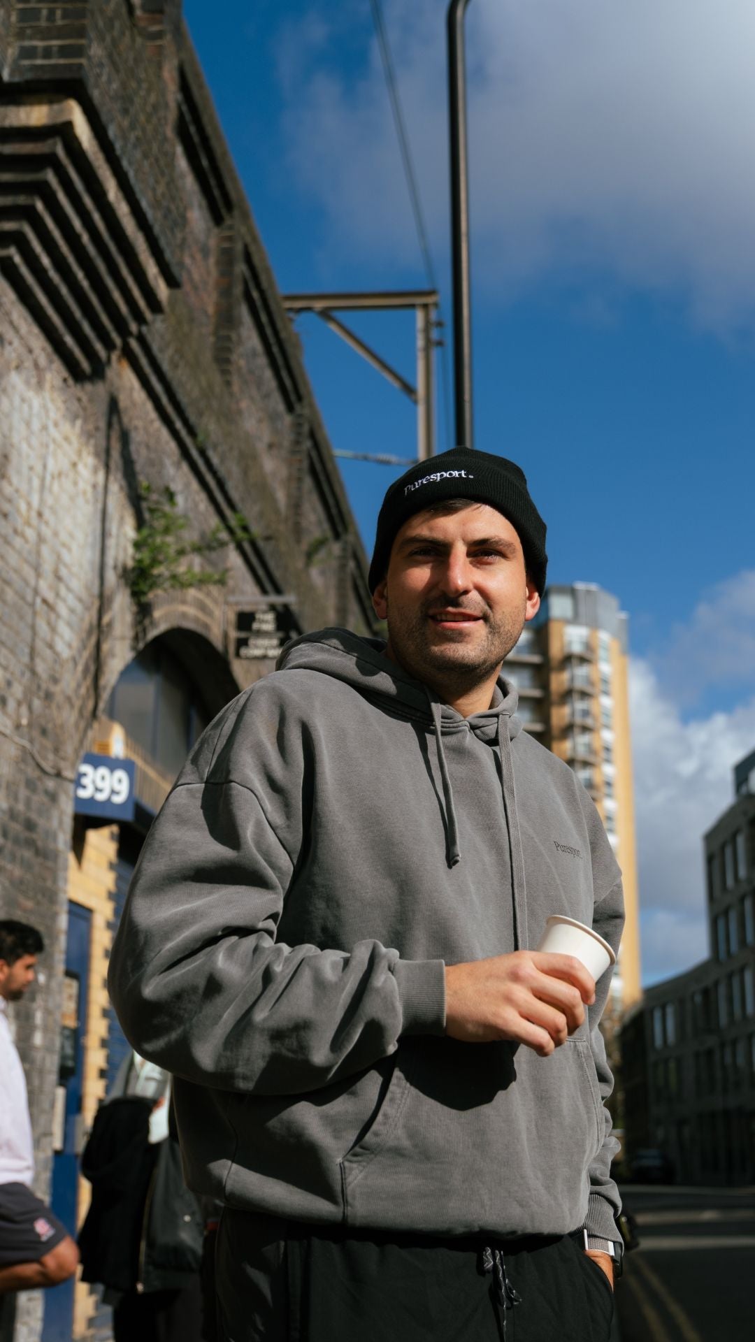 Runner in a grey hoodie holding a coffee cup outdoors with buildings and blue sky in the background. 