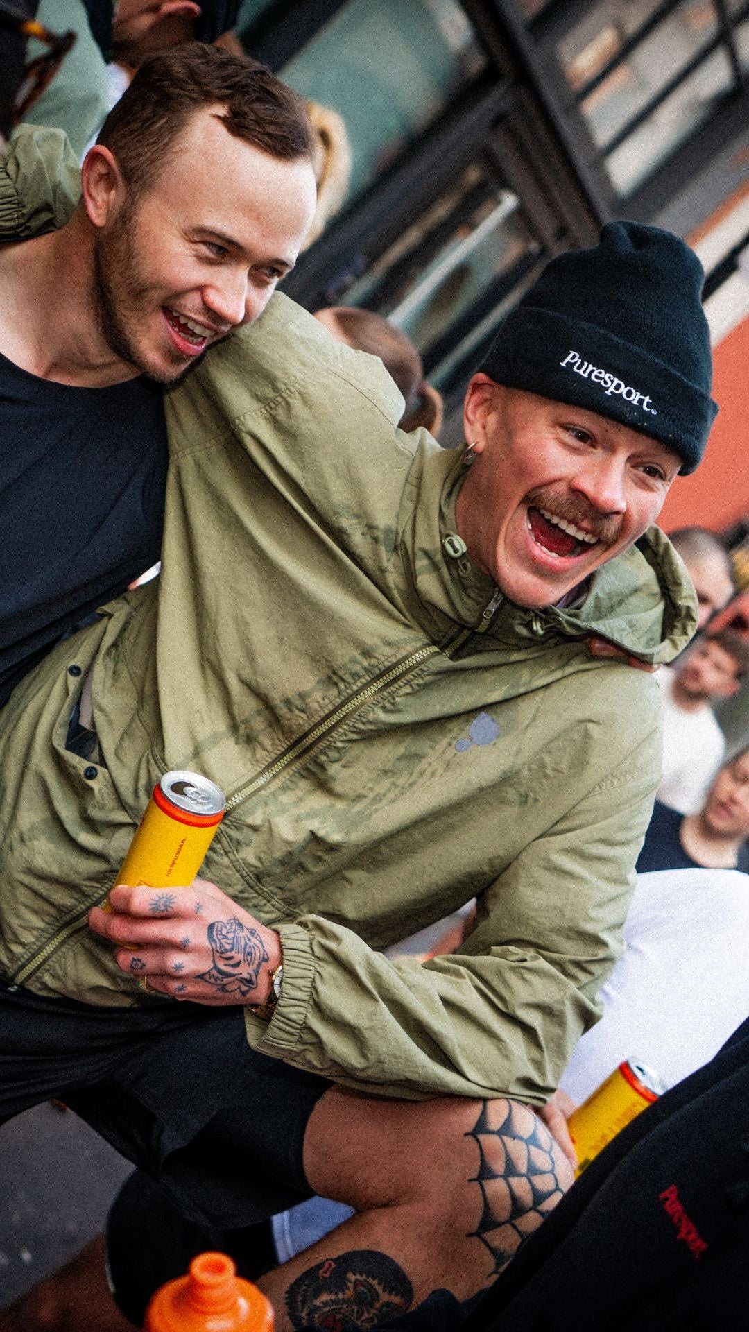Two men laughing together, one wearing a green jacket by Puresport and black cap with 'Puresport' branding.