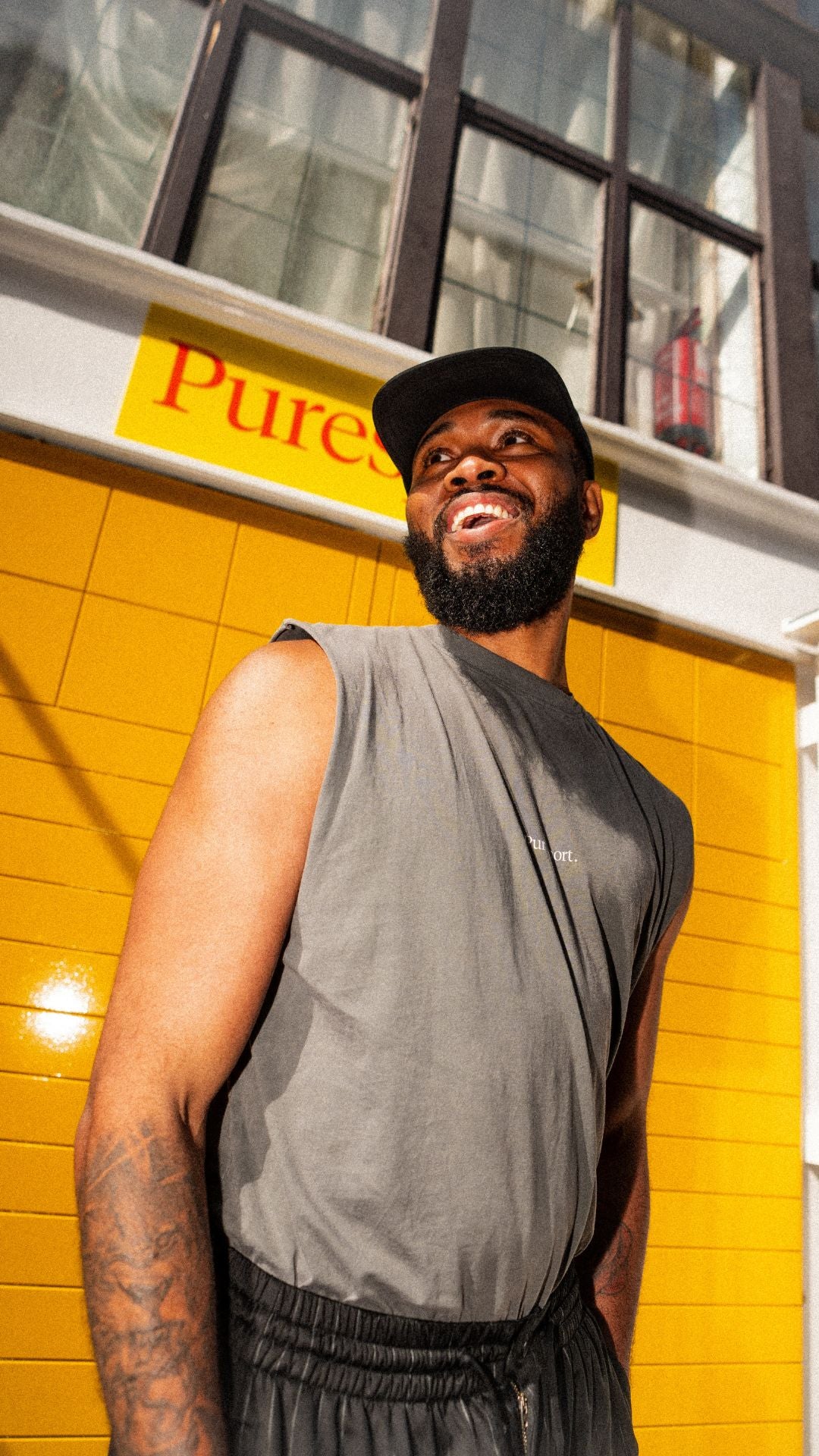 Man in grey sleeveless shirt and black cap standing against a yellow wall with Puresport sign.