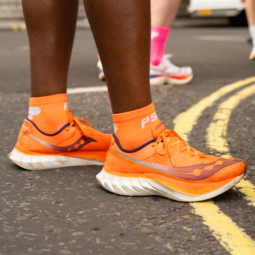 Person wearing orange running shoes with Puresport running socks on a road with blurred background.