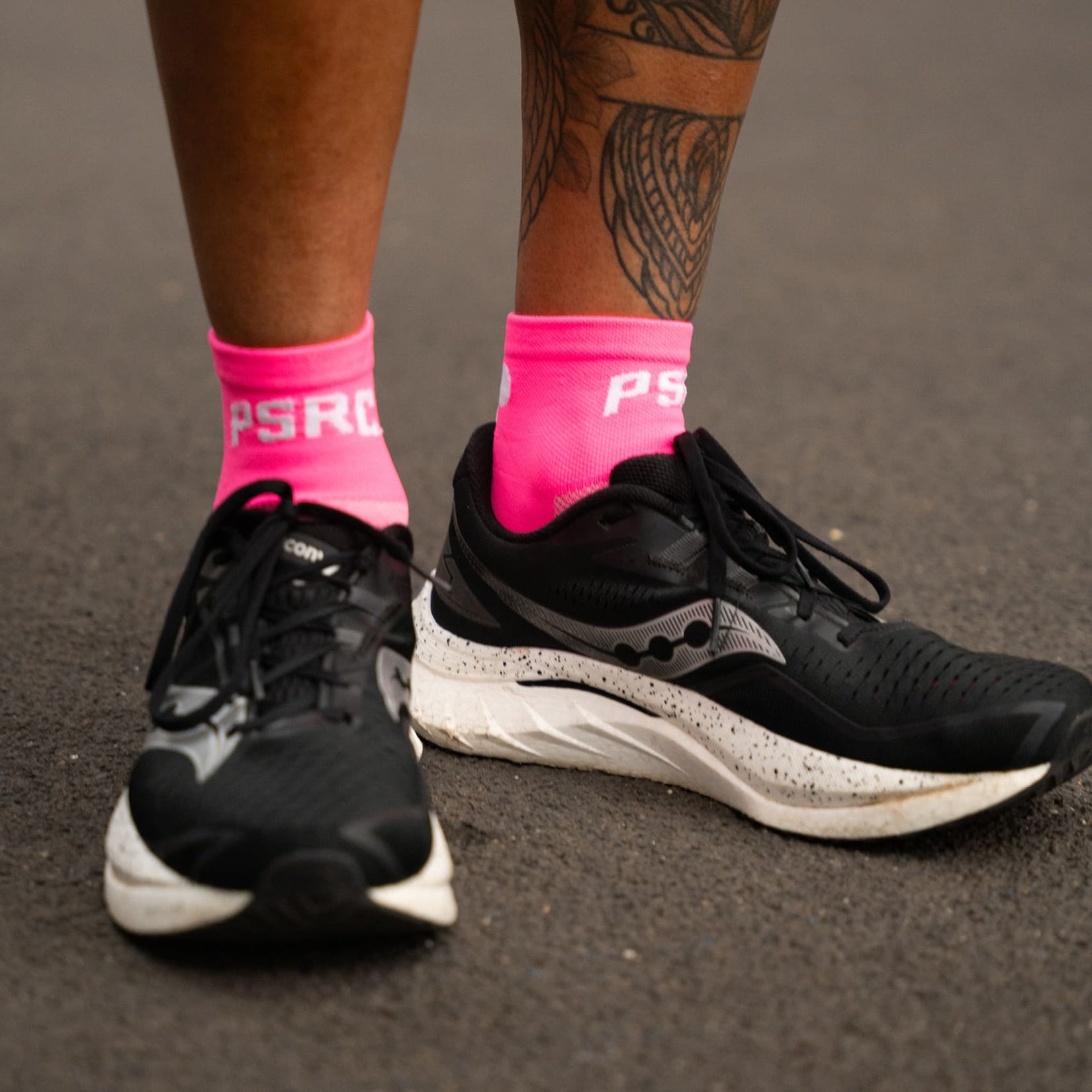 Black running shoes with white soles worn on a gray surface, featuring pink socks with 'PSRC' branding.