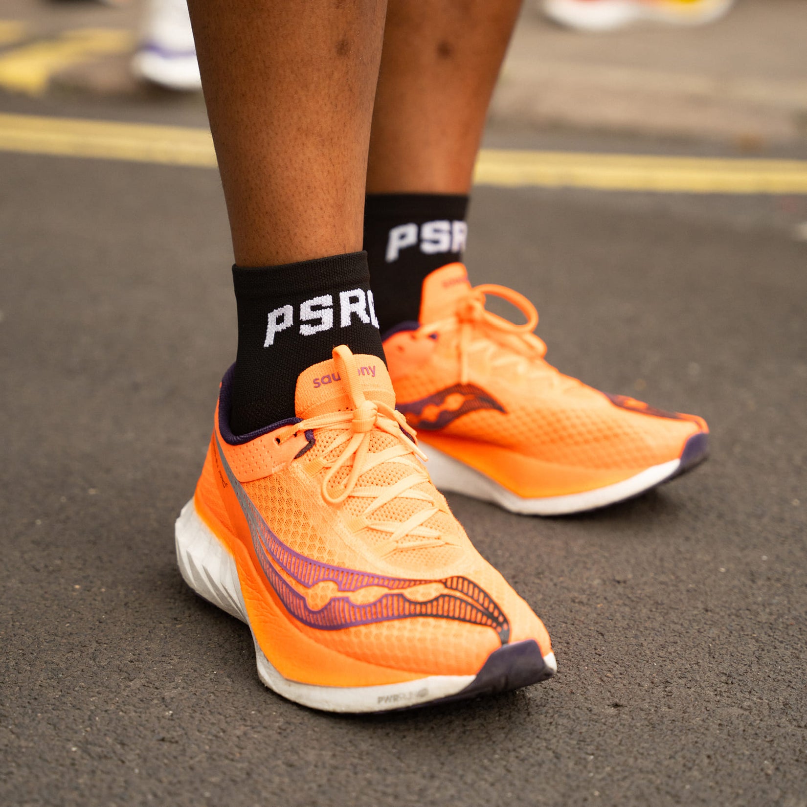 Orange running shoes with black socks featuring 'PSRC' brand on a road background