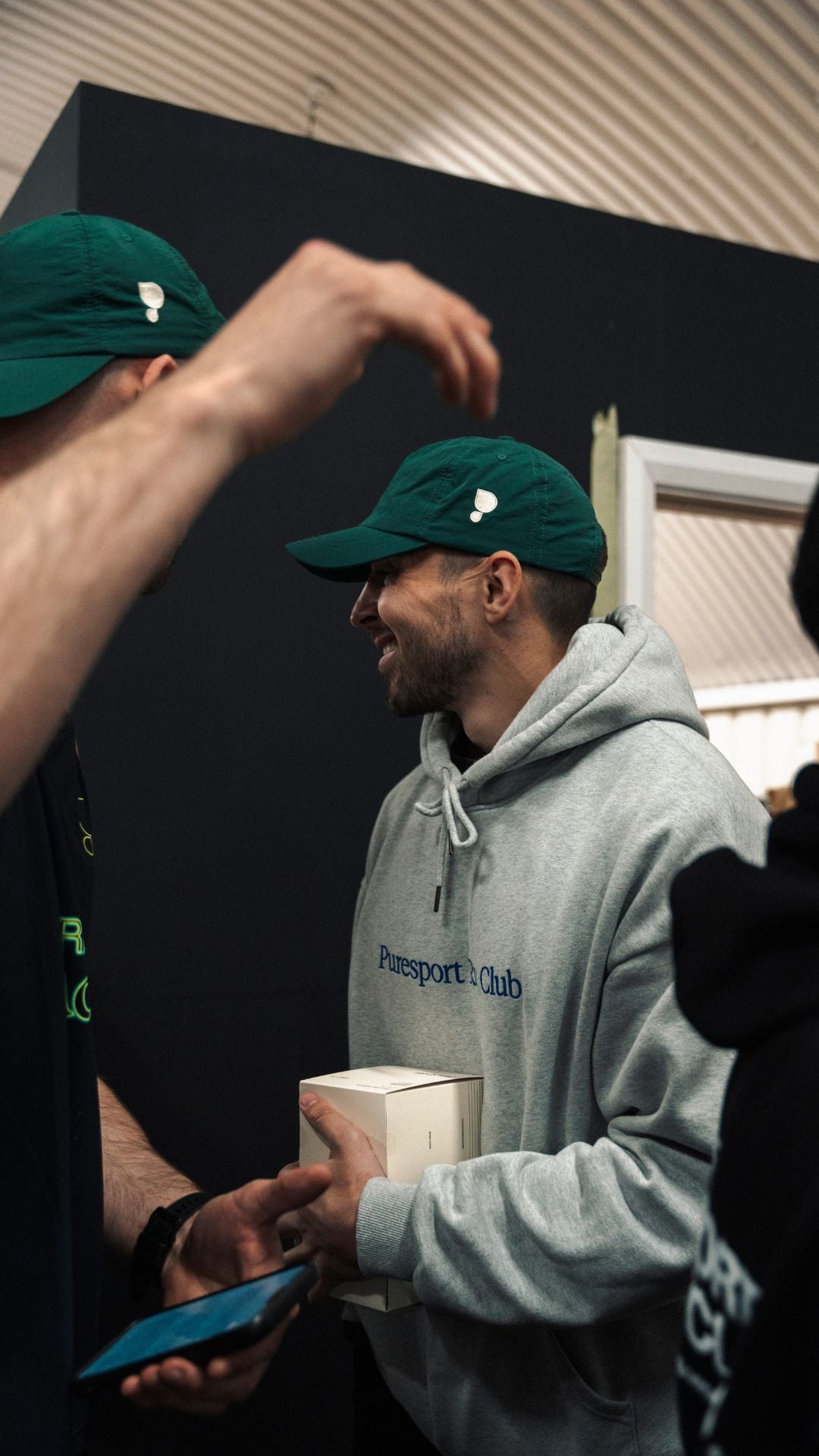 Male wearing a green cap by Puresport and grey hoodie with 'Puresport Running Club' branding, interacting with another person.