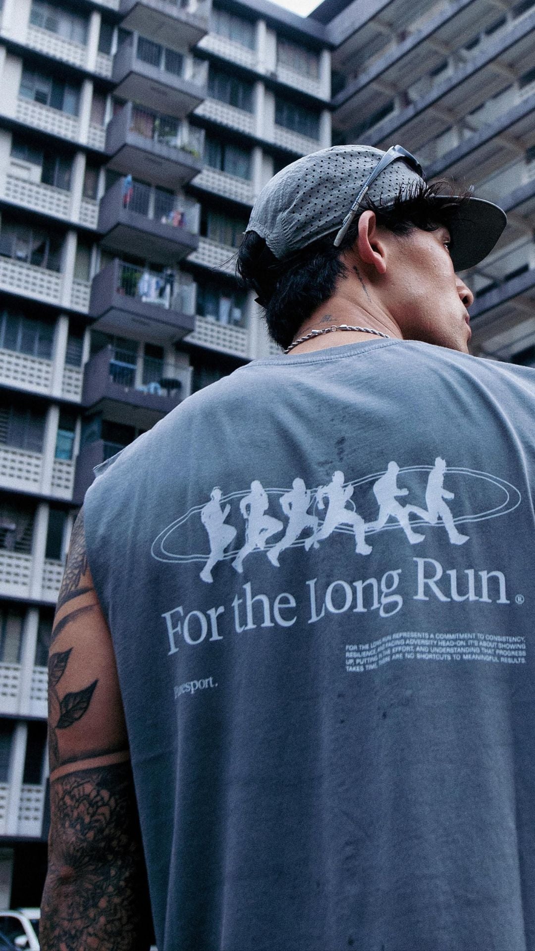 Runner wearing a gray cut off with 'For the Long Run' text and graphic, standing in front of a building.