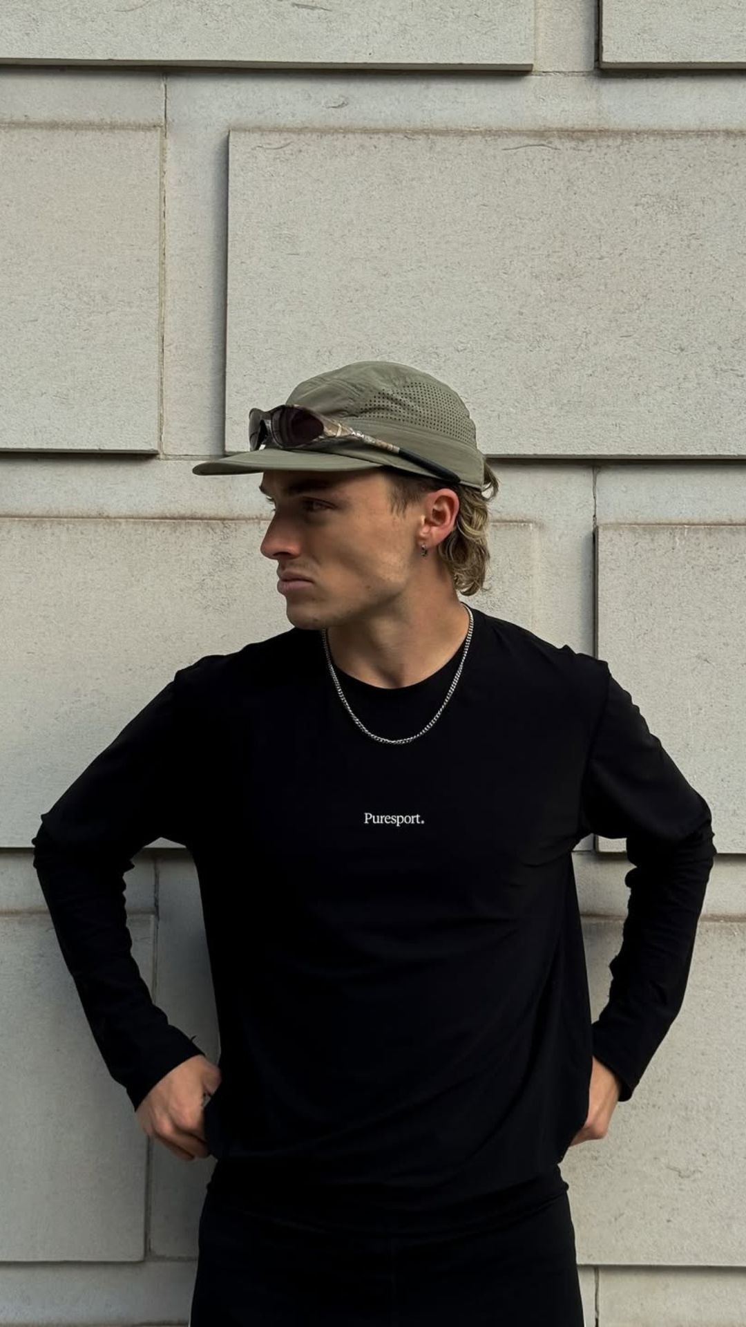 A runner wearing a black long-sleeve shirt by Puresport and a run cap, standing against a light-coloured wall.