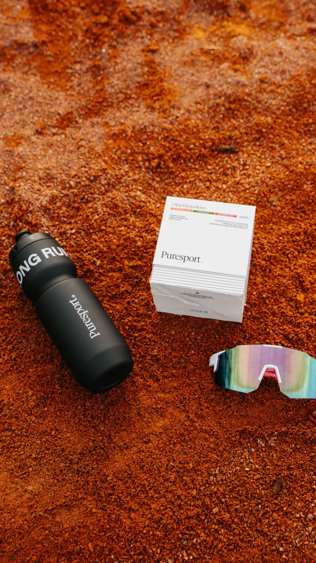Black water bottle by Puresport, box of Electrolytes and colourful running sunglasses on a rust-colorued surface