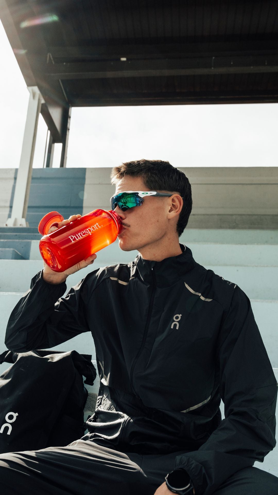 Runner in black jacket drinking from a red nalgene bottle with oakleys on, sitting outdoors.