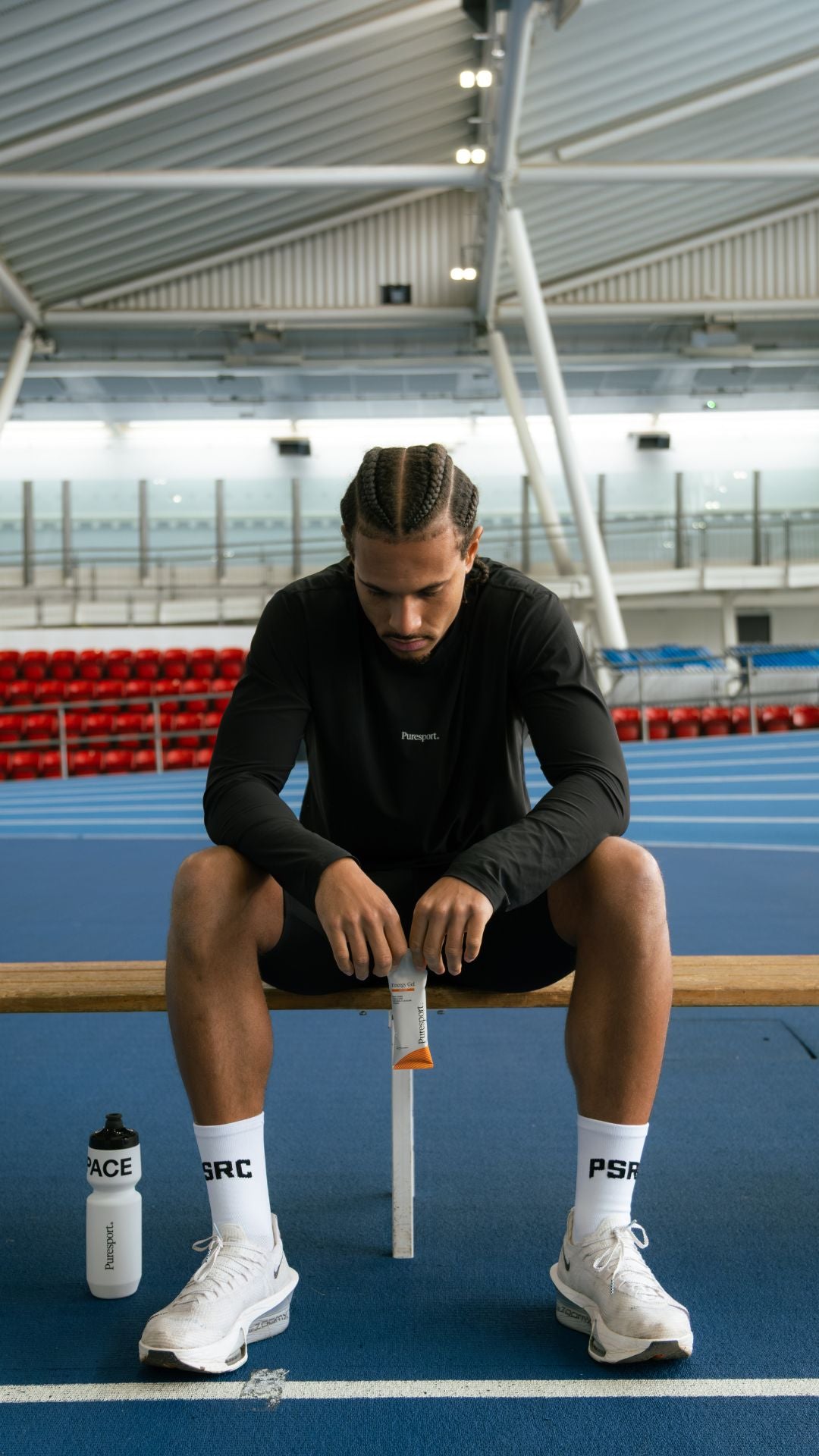 Person sitting on a track and field with sports equipment, wearing black athletic wear and white shoes holding an energy gel. 